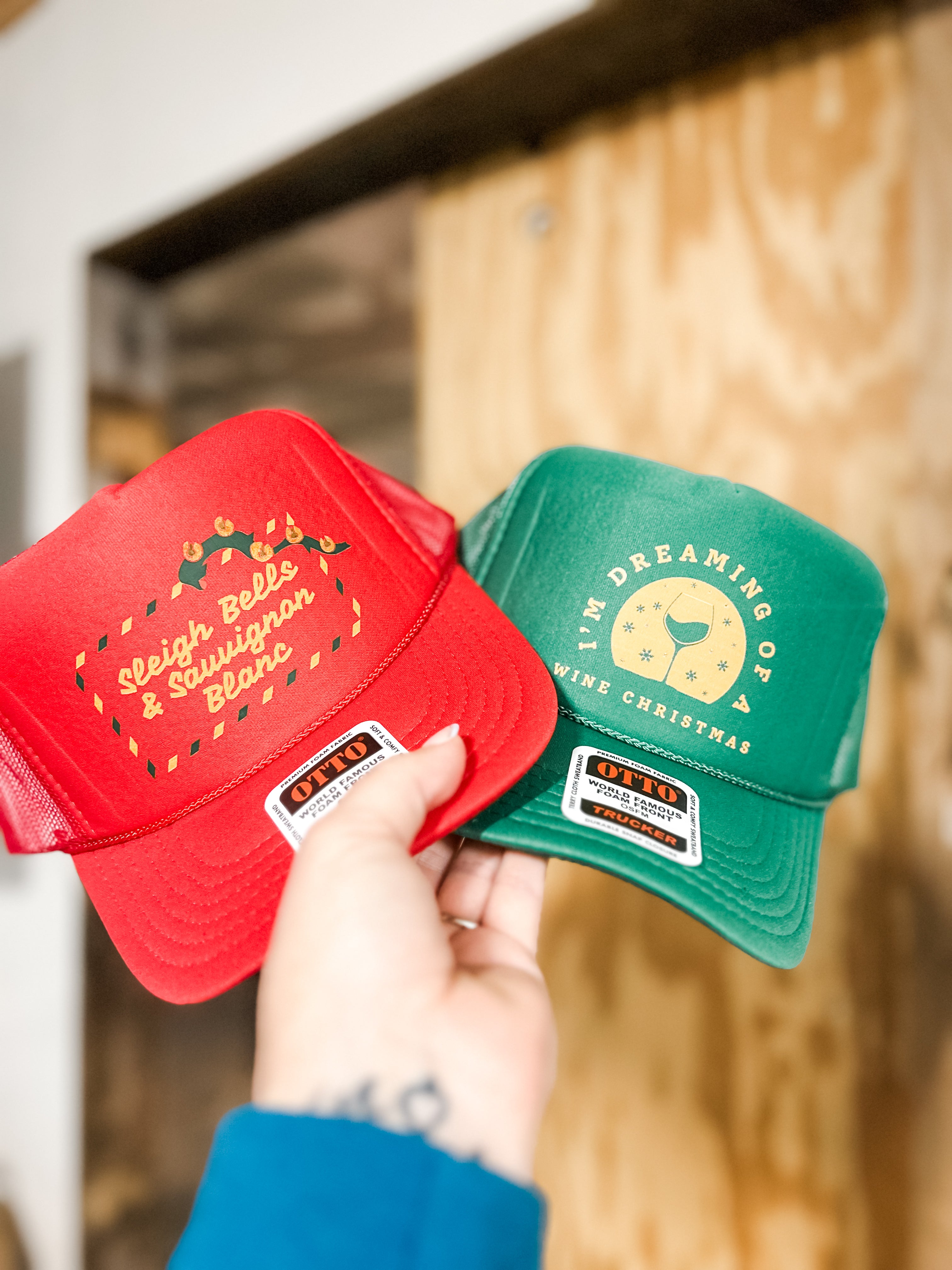Red and green Christmas-themed hats for wine lovers held by a hand that say im dreaming of a wine christmas and sleigh bells and sauvignon blanc from corkie apparel